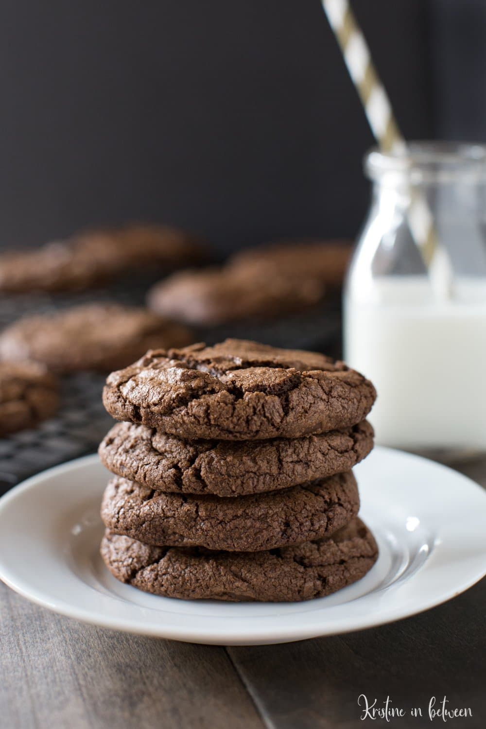 Dark chocolate cookies sitting on a white plate with a jar of milk next to them.