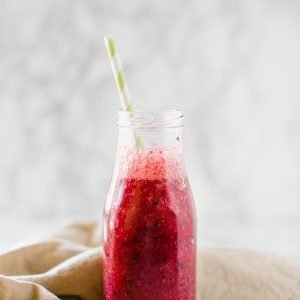 A mixed berry smoothie in a glass milk bottle with a brown napkin behind it.