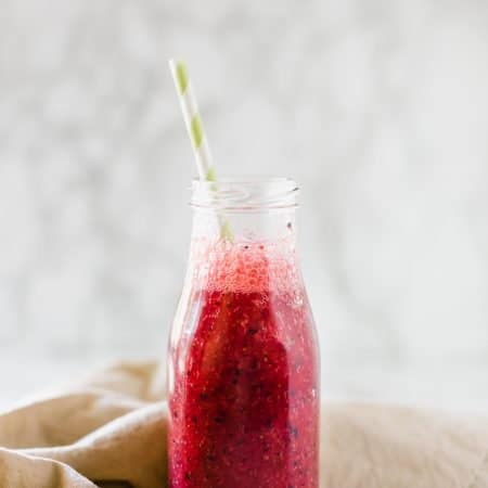 A mixed berry smoothie in a glass milk bottle with a brown napkin behind it.