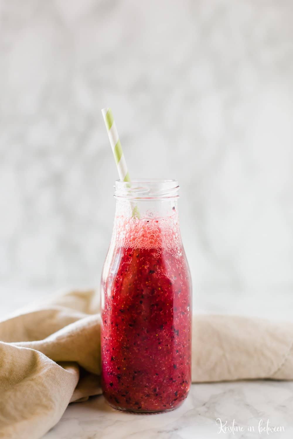 A mixed berry smoothie in a glass milk bottle with a brown napkin behind it.