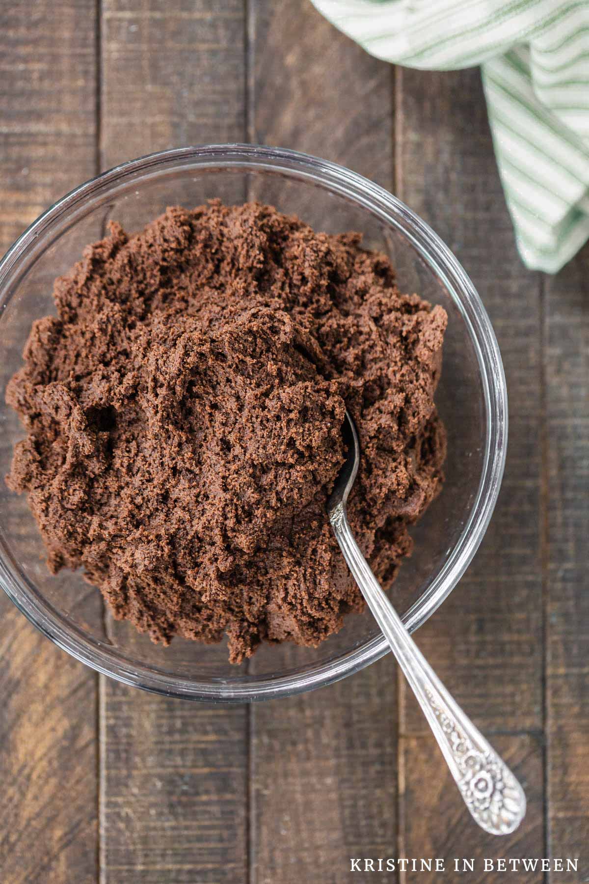 A glass bowl of chocolate cookie dough with a spoon in it.