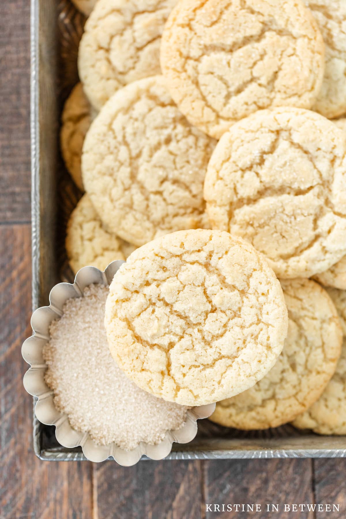Sugar cookies stacked up in a tin with a small bowl of sugar sitting next to them.