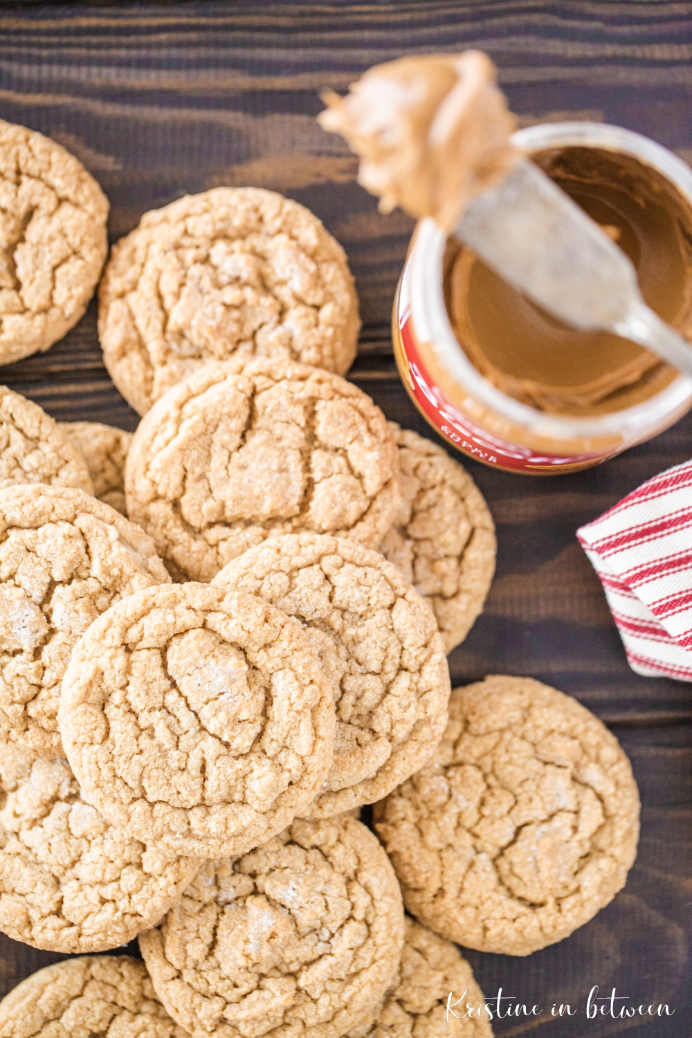 Cookies stacked up on a table with a jar of cookie butter, a knife, and a red and white striped napkin.