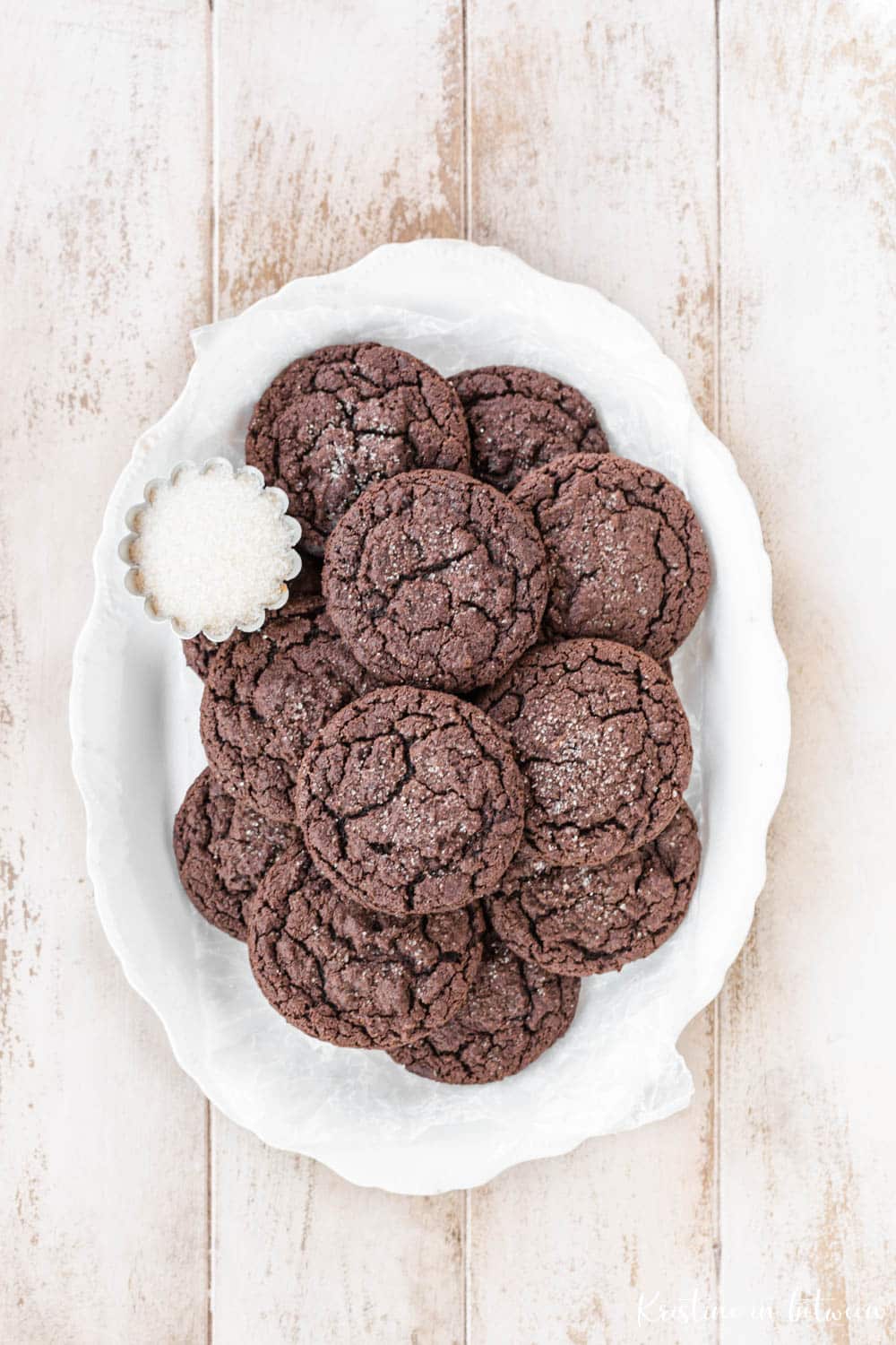 Chocolate cookies stacked up on a white platter with a small bowl of sugar.