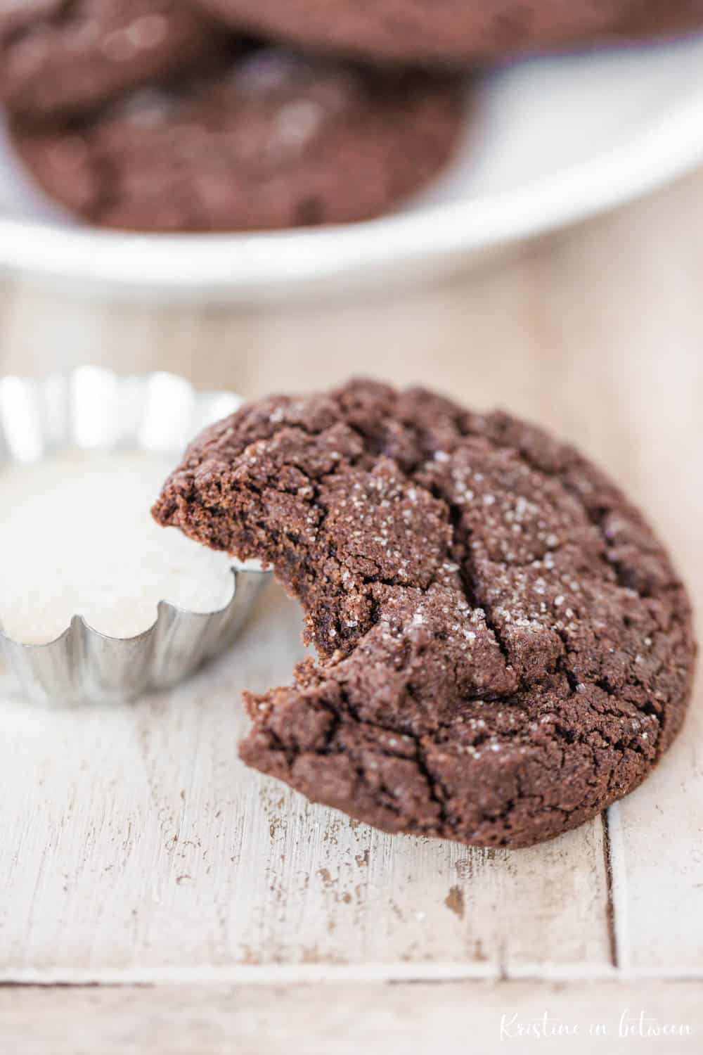 A chocolate cookie with a bite out of it leaning on a small bowl of sugar.