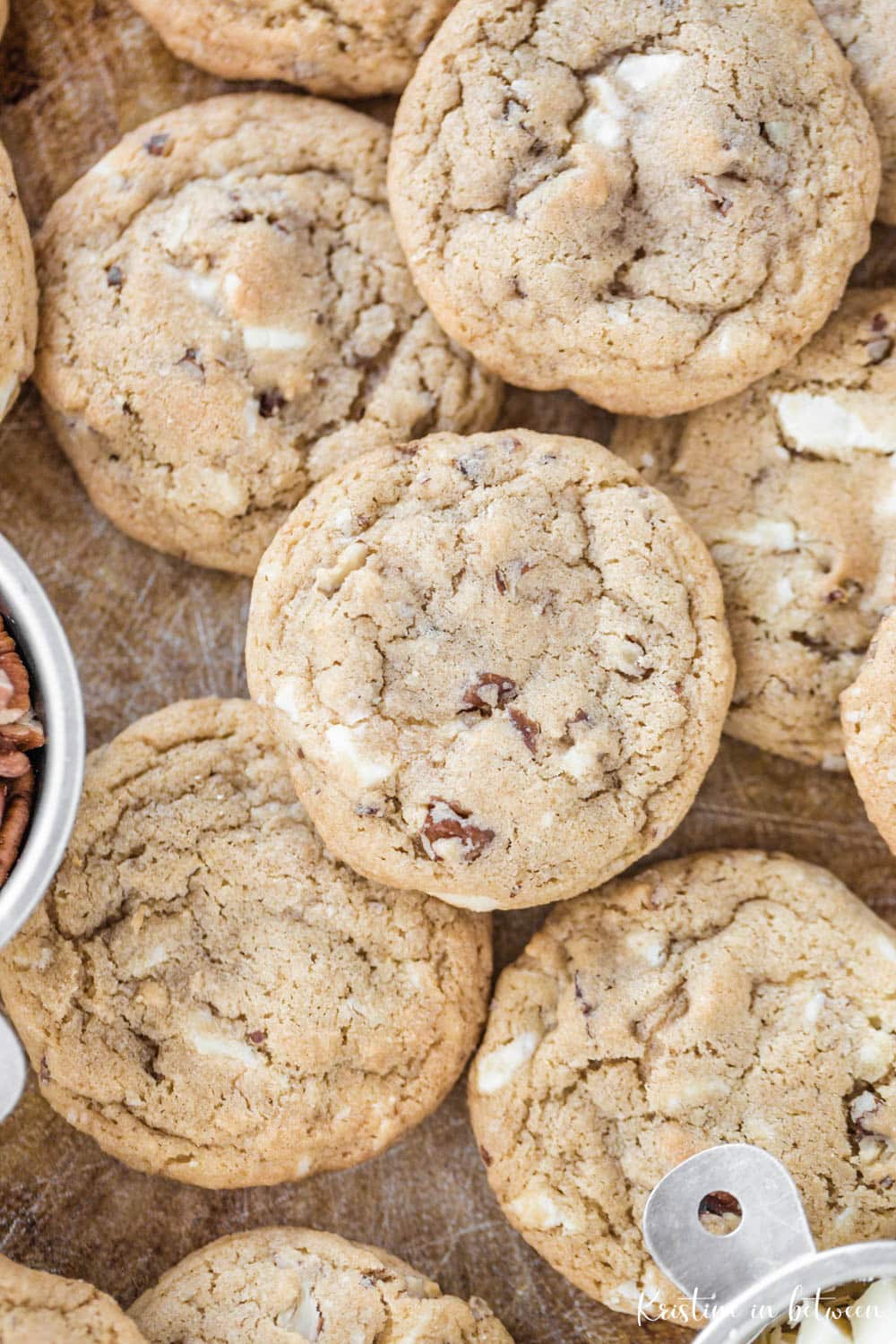 Cookies laying on a cookie sheet with a small measuring cup full of pecans and another full of chopped white chocolate.