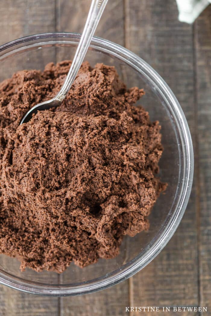 A glass bowl of chocolate cookie dough with a spoon in it.