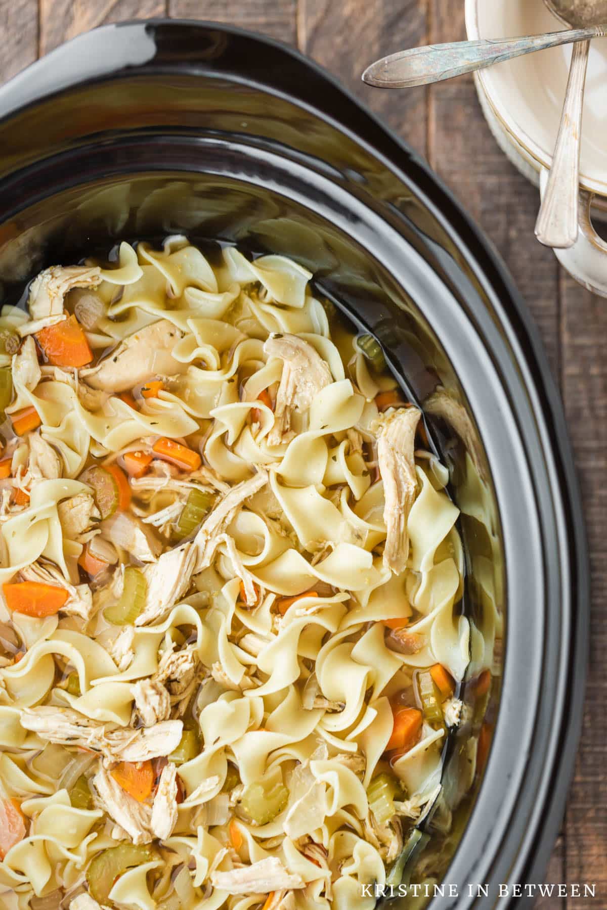 Chicken noodle soup in the crock of a Crock-pot with two bowls and two spoons sitting next to it.