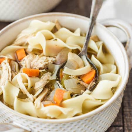 Two bowls of chicken noodle soup with antique spoons and an orange striped napkin in the background.