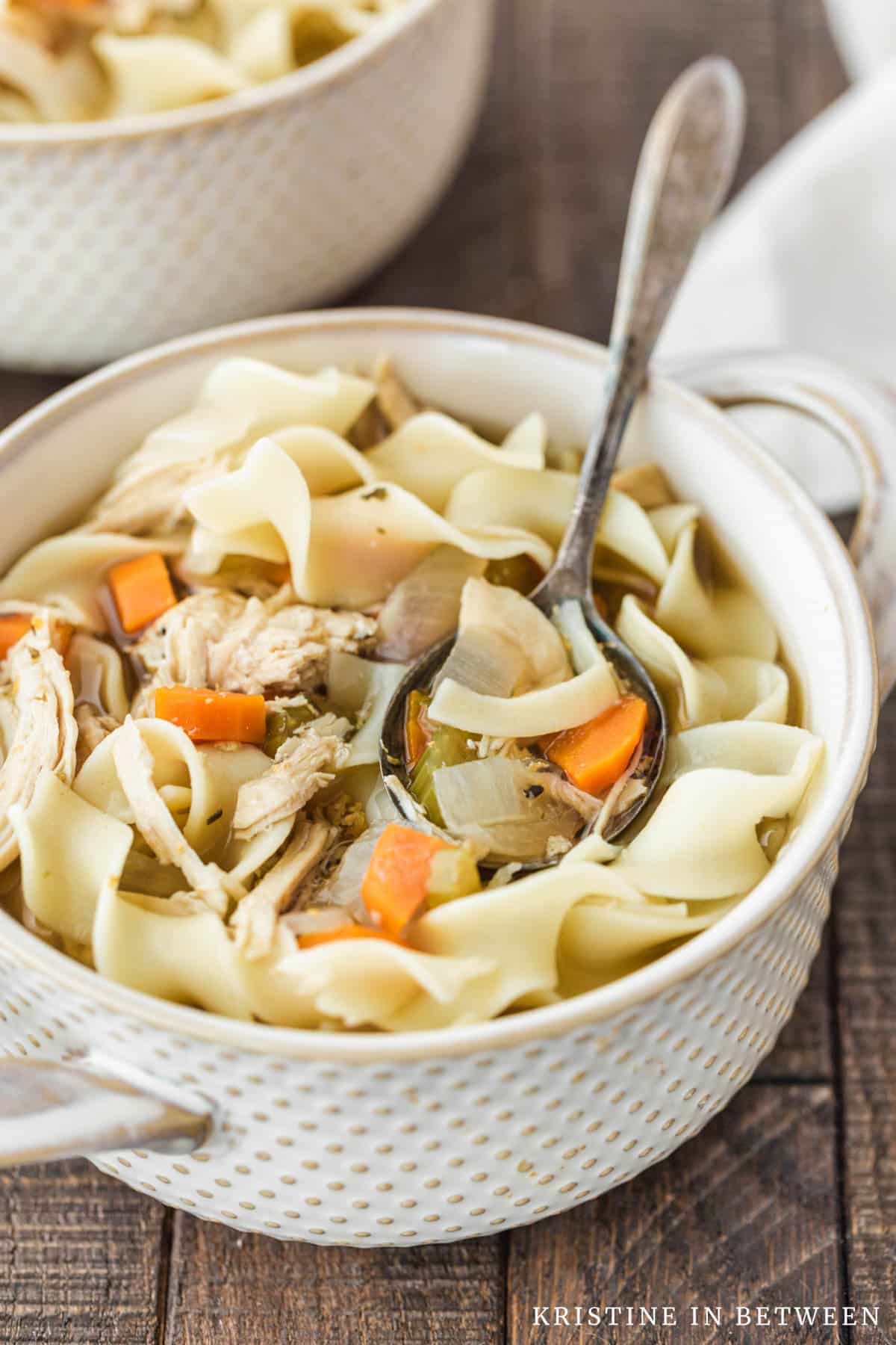 Two bowls of chicken noodle soup with antique spoons and an orange striped napkin in the background.