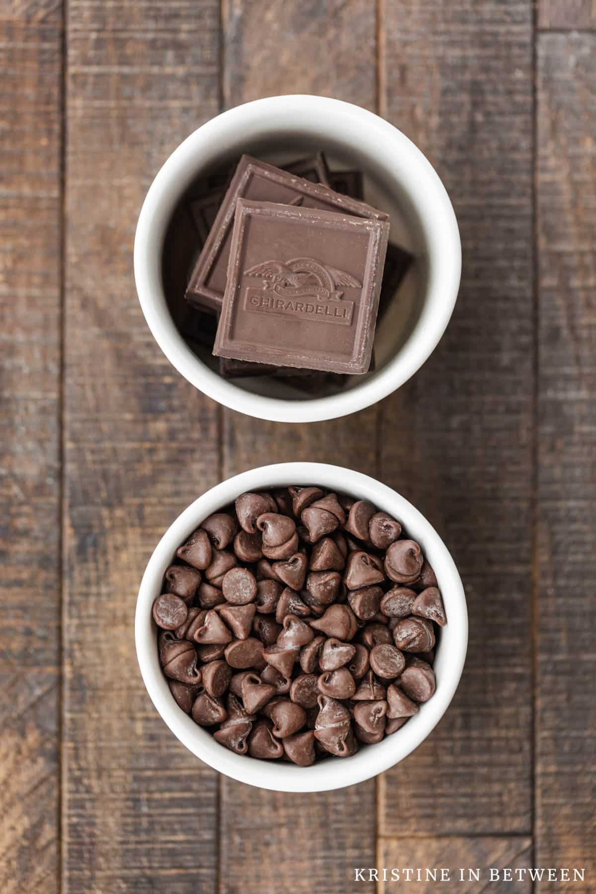 Two bowls of chocolate sitting on a wooden table, one with baking chocolate and one with chocolate chips.
