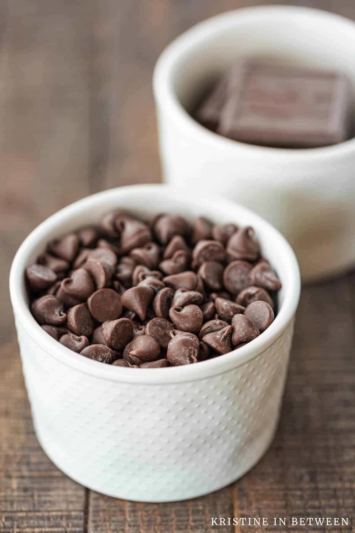 Two bowls of chocolate sitting on a wooden table, one with baking chocolate and one with chocolate chips.