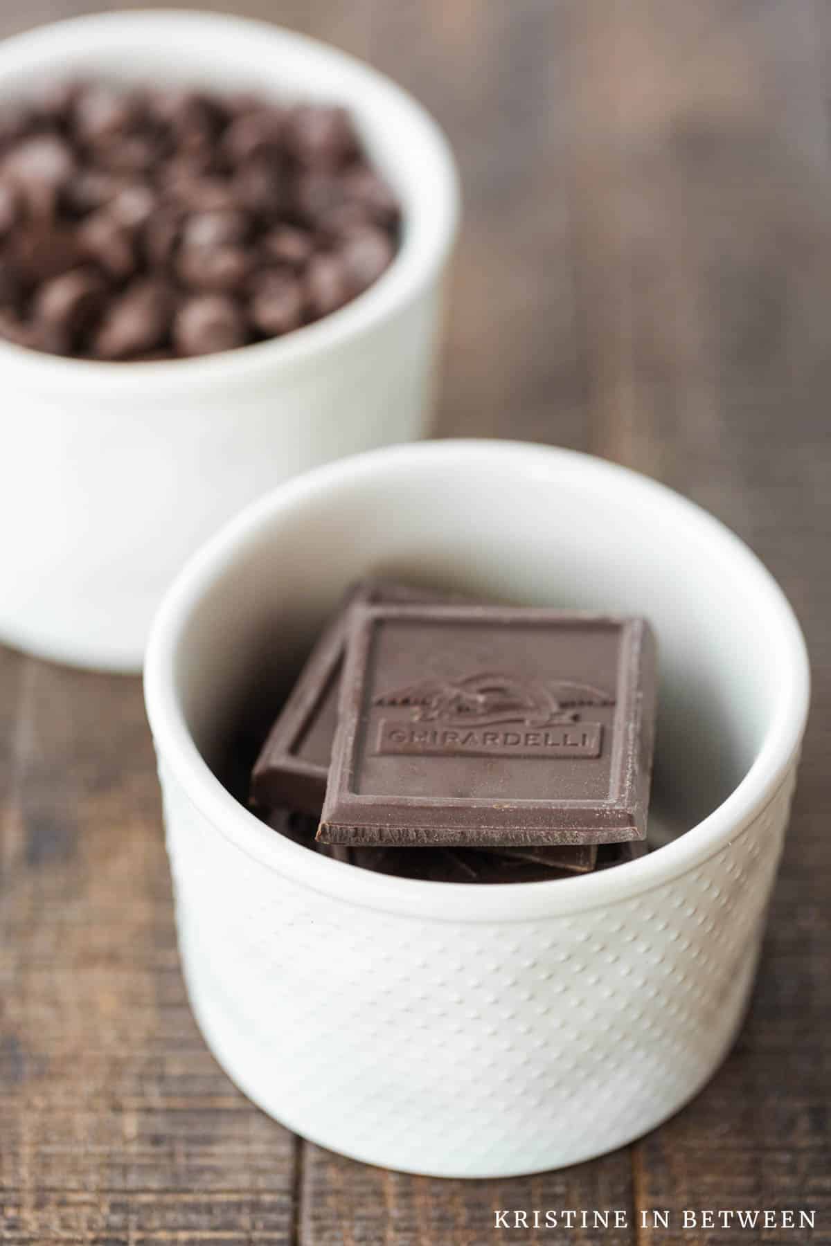 Two bowls of chocolate sitting on a wooden table, one with baking chocolate and one with chocolate chips.