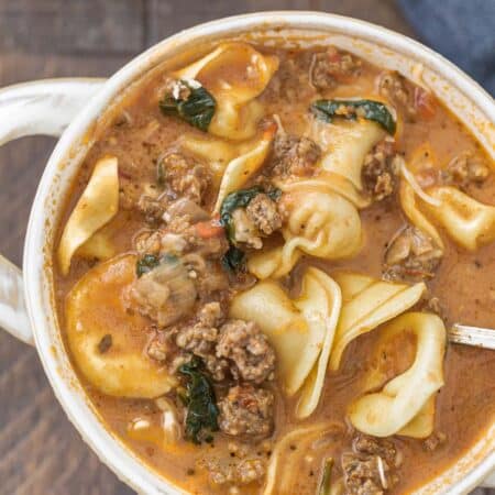 A bowl of sausage tortellini soup with a spoon and a blue napkin laying next to it.