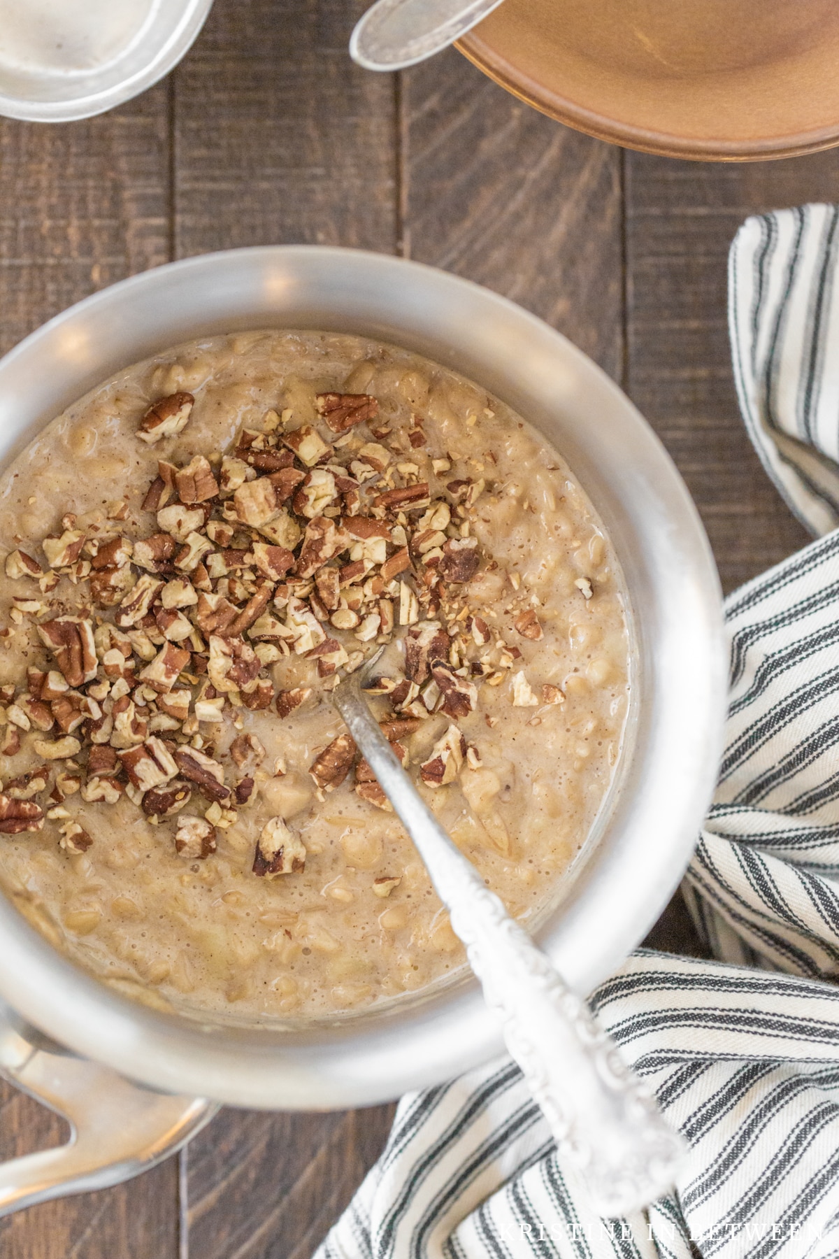 Cooked oatmeal sitting in a pan with a spoon and bowls in the background.