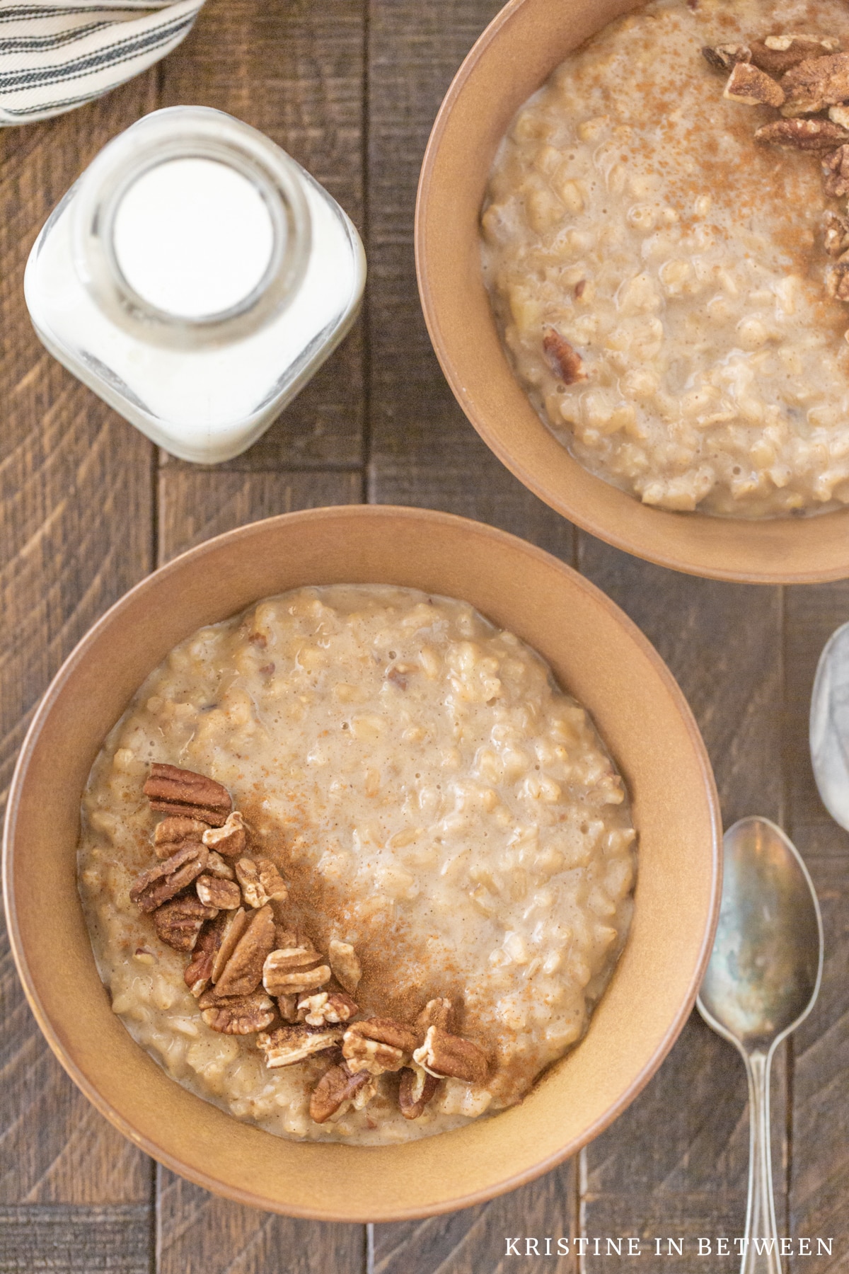 Two bowls of banana pecan oatmeal sitting with spoons and a small jar of milk in the background.