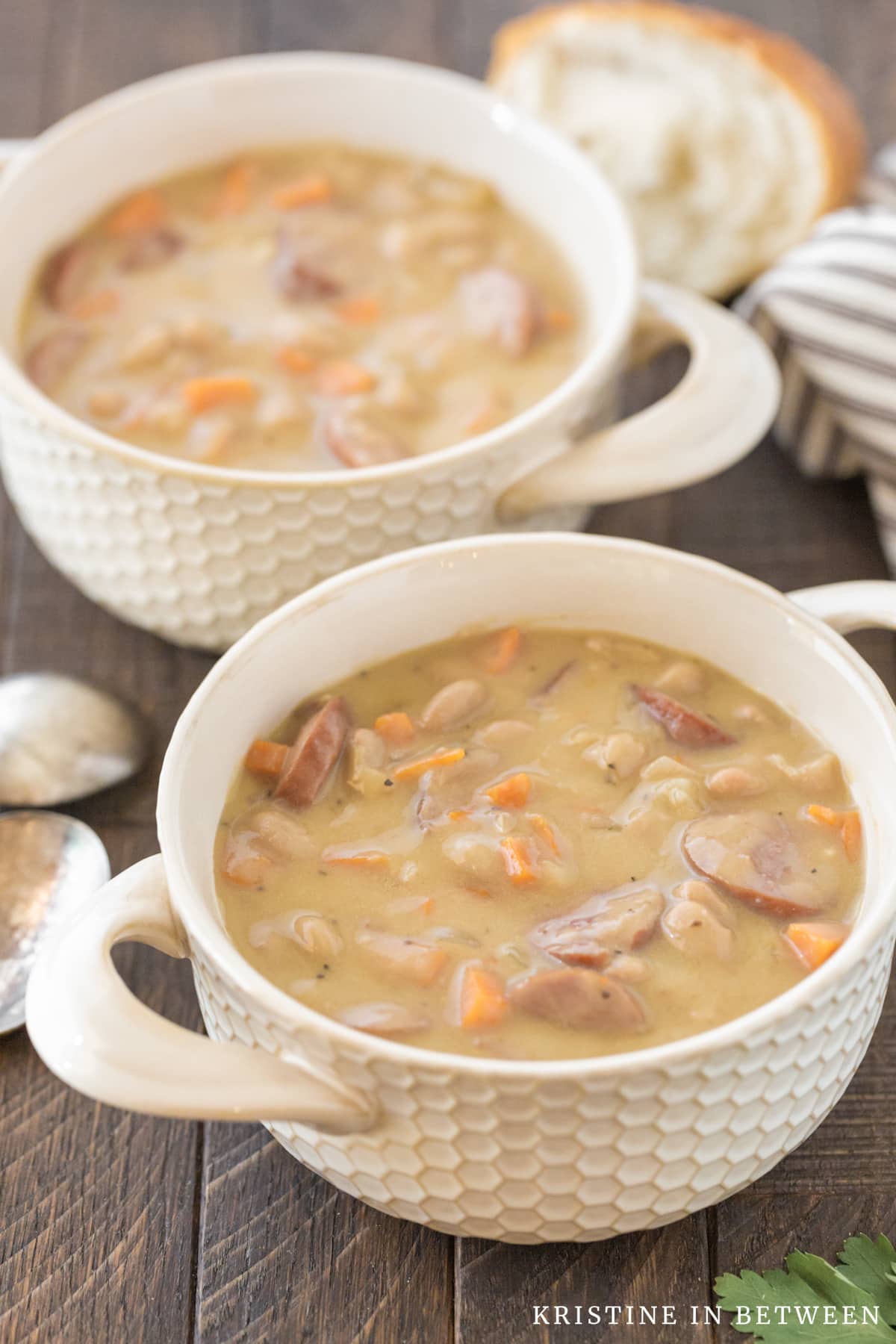Two bowls of kielbasa soup sitting two spoons and a piece of bread.
