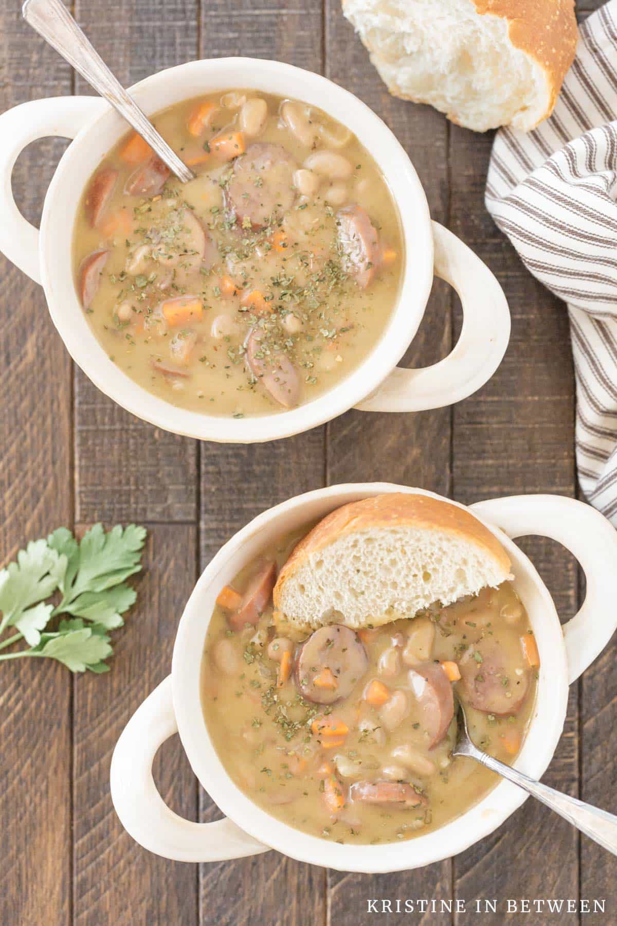 Two bowls of kielbasa and white bean soup sitting with a piece of bread and two spoons.
