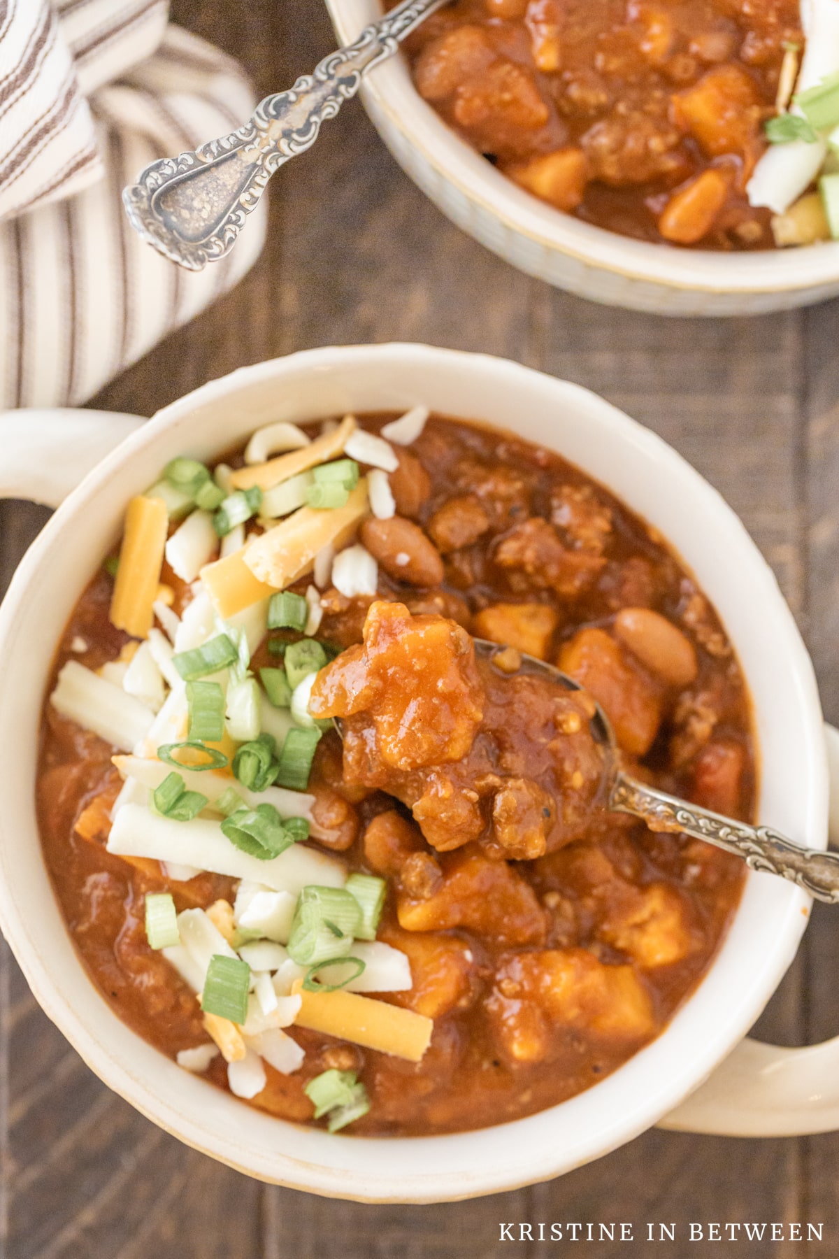 Two bowls of sweet potato chili with spoons in them, topped with cheese and green onion.