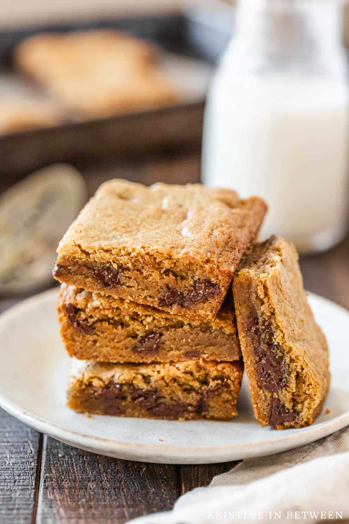 Chocolate chip cookie bars stacked up on a white plate with a jar of milk in the background.