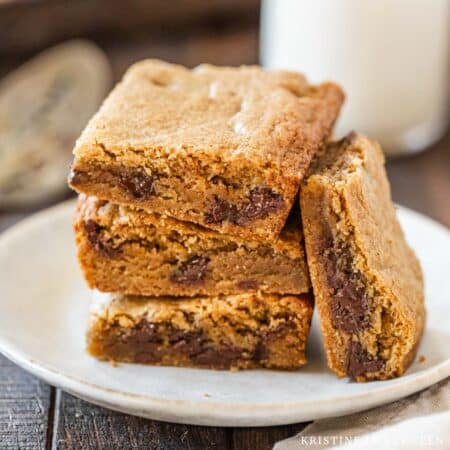 Chocolate chip cookie bars stacked up on a white plate with a jar of milk in the background.