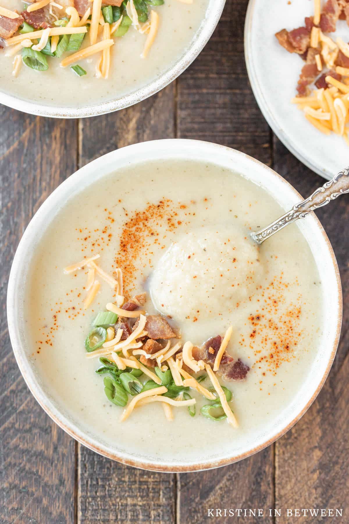 A bowl of cauliflower soup with a spoon in it, garnished with paprika, green onion, bacon bits, and cheese.