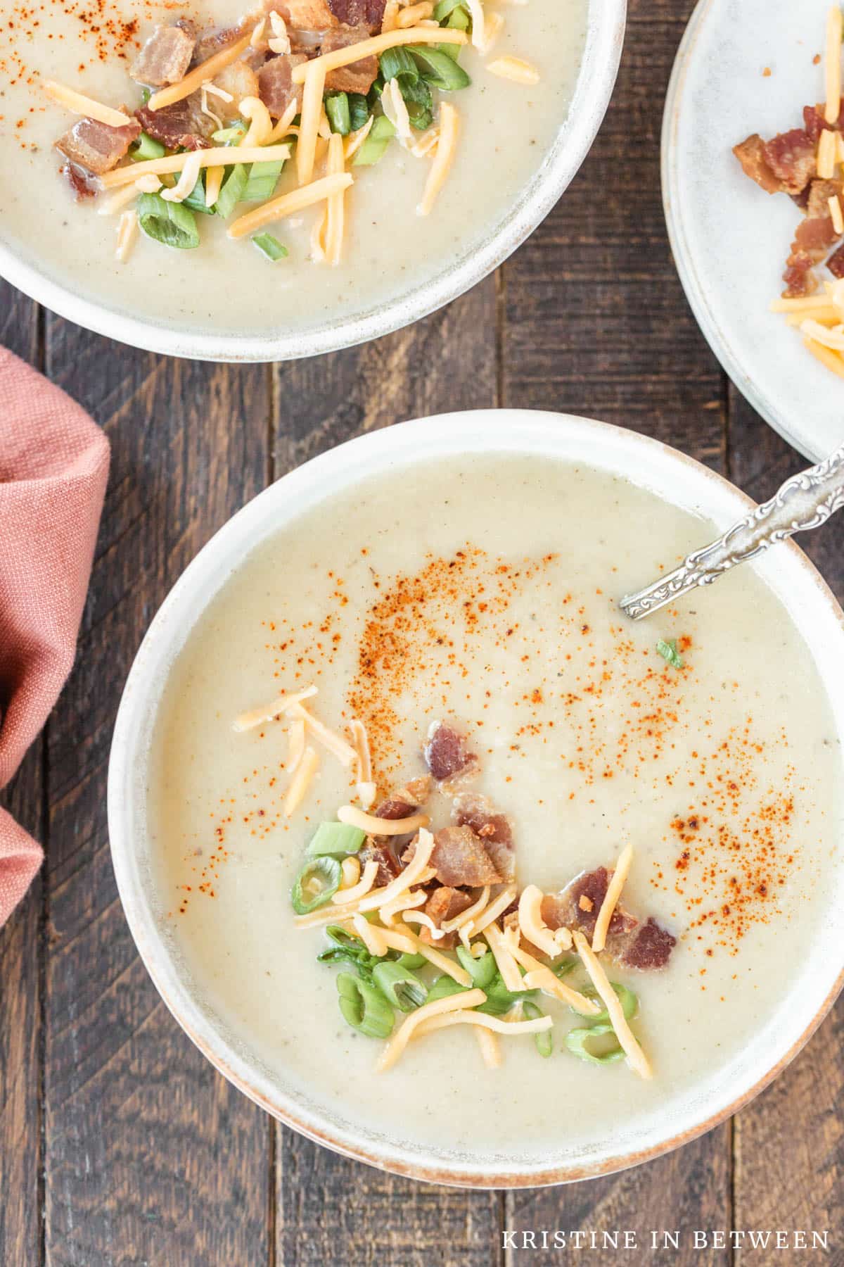 A bowl of cauliflower soup with a spoon in it, garnished with cheese, green onion, and bacon bits.