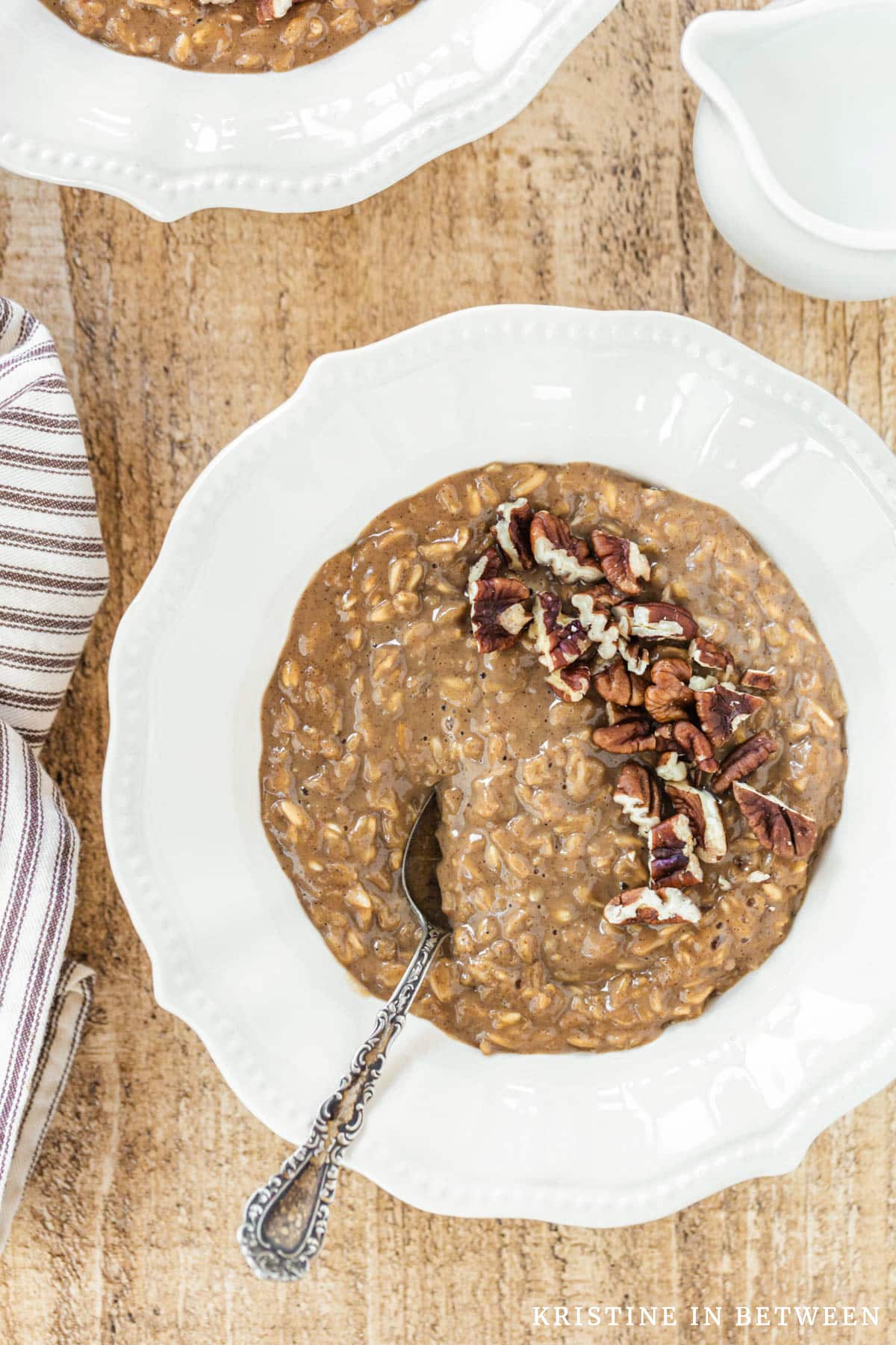 A bowl of gingerbread oatmeal topped with chopped pecans sitting with a spoon and a small pitcher of milk.