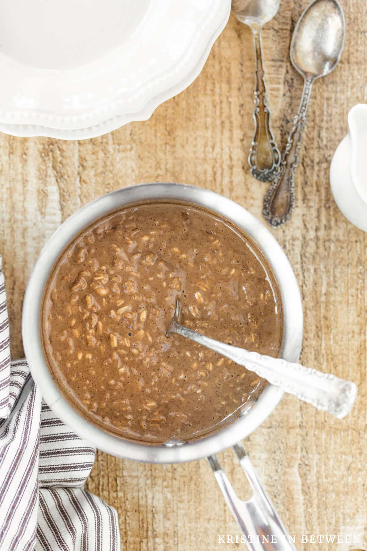 Cooked oatmeal in a stainless steel pan with two bowls and spoons in the background.