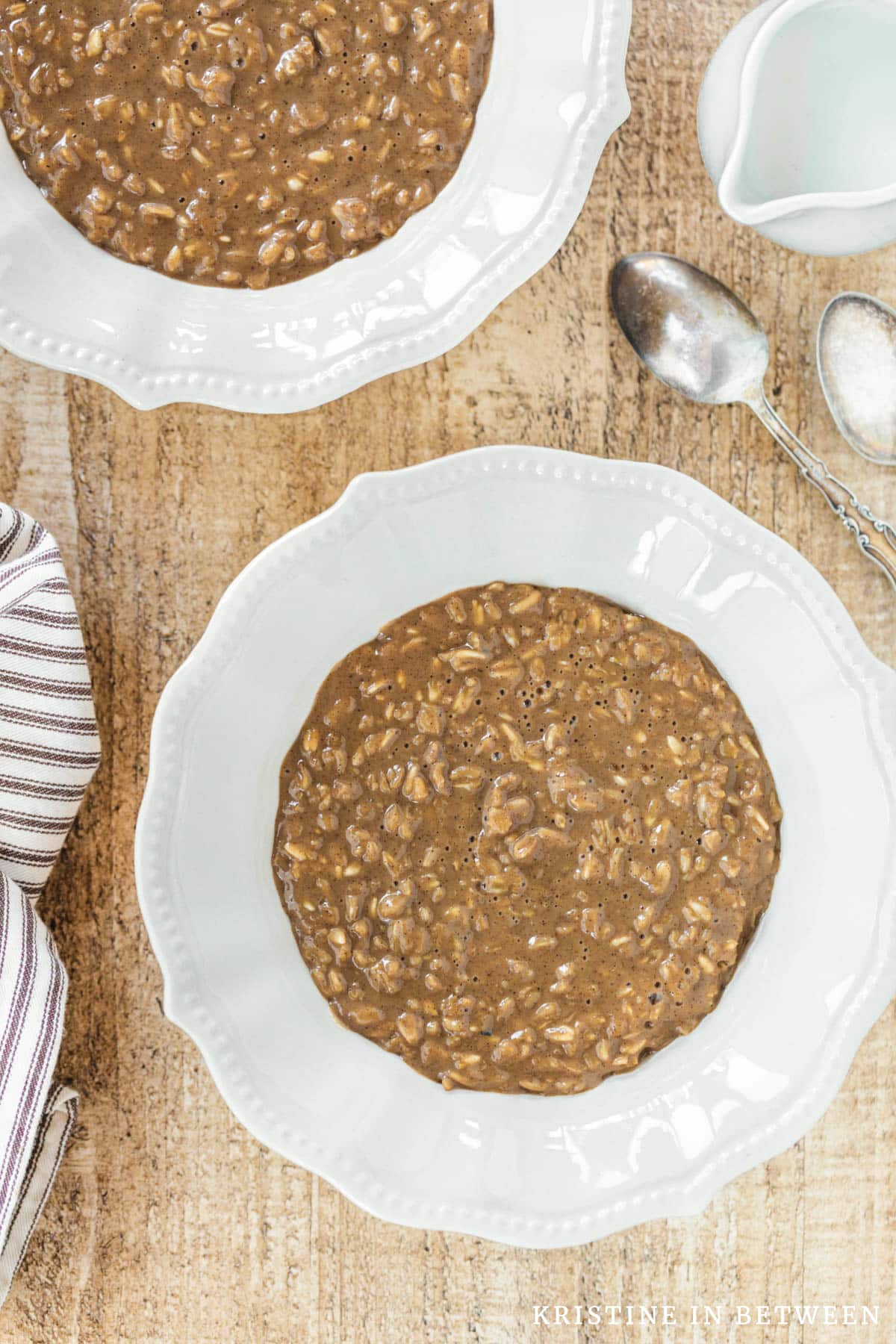 Two bowls of cooked oatmeal sitting with two spoons and a small pitcher of milk.