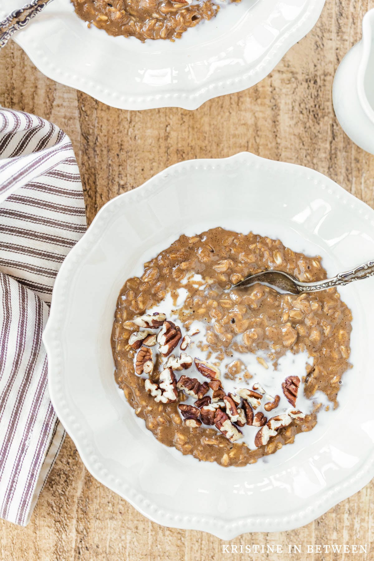 A bowl of gingerbread oatmeal topped with chopped pecans sitting with a spoon and a small pitcher of milk.