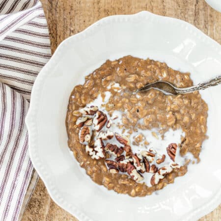 A bowl of gingerbread oatmeal topped with chopped pecans sitting with a spoon and a small pitcher of milk.