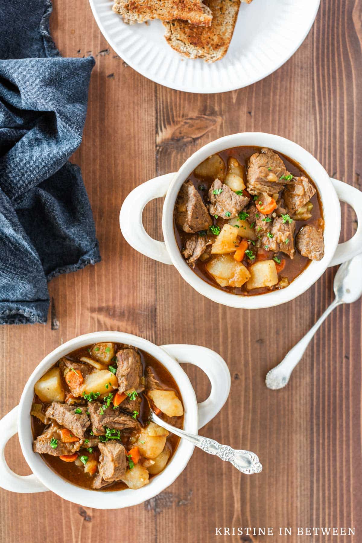Two bowls of beef stew with spoons and a plate with toasted brown bread next to them.