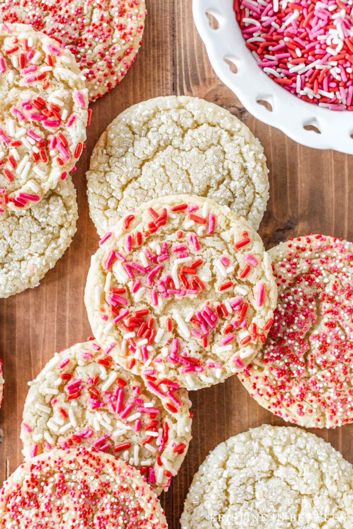 Valentine sugar cookies with red, white, and pink sprinkles laying on a wooden board with a small bowl of sprinkles.