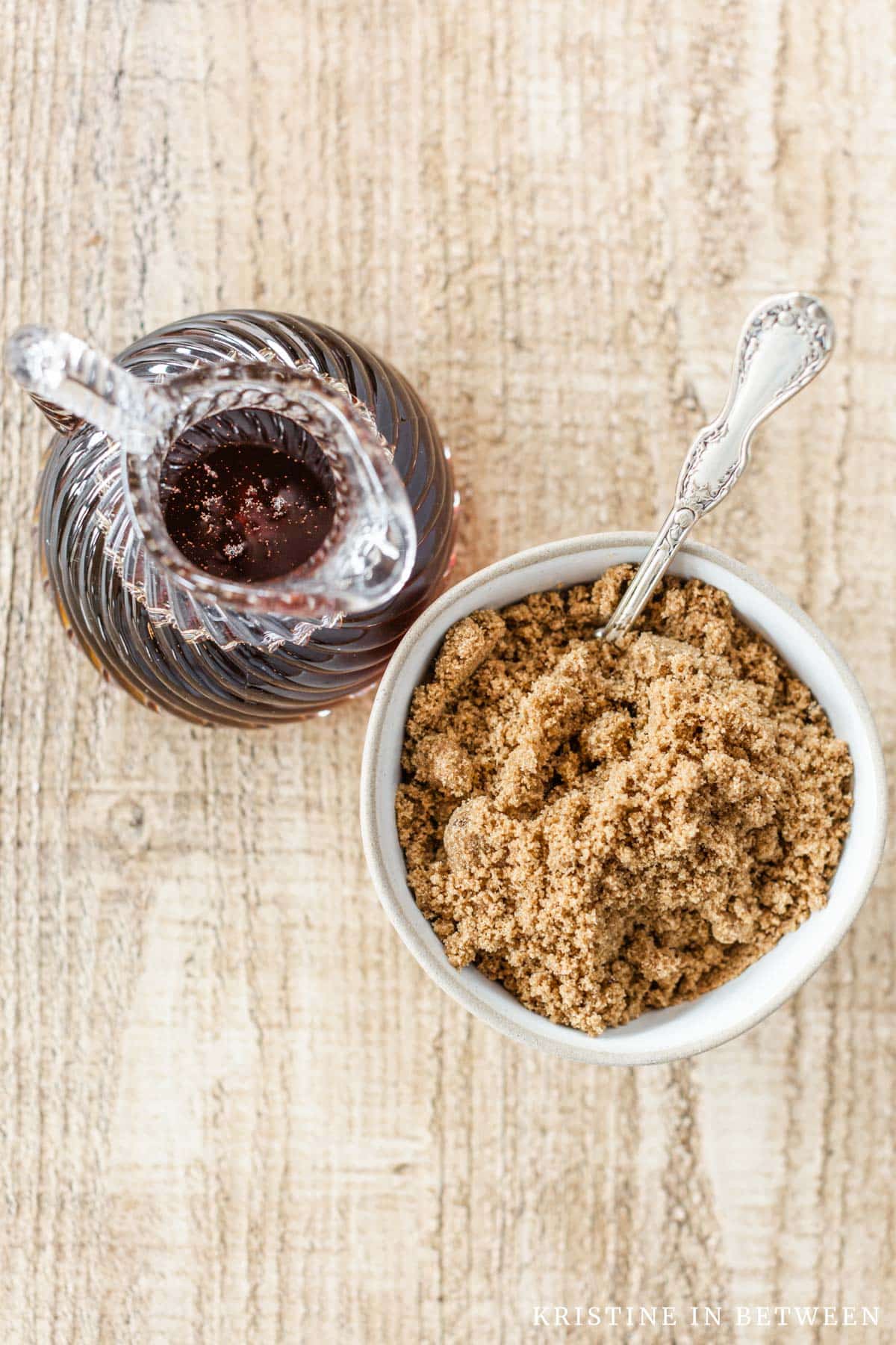 A small bowl of brown sugar with a spoon in it sitting with a jar of maple syrup.