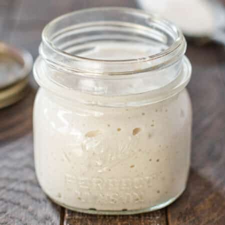 A sourdough start in a glass jar with a scoop of flour in the background.
