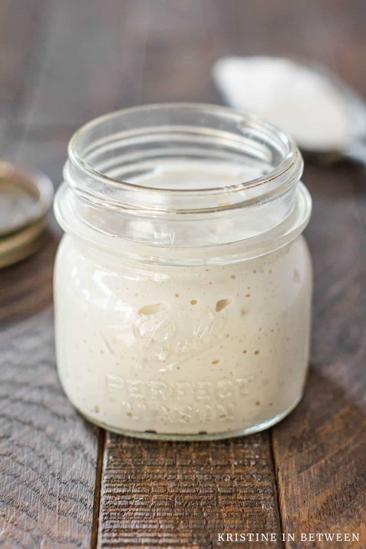 A sourdough start in a glass jar with a scoop of flour in the background.