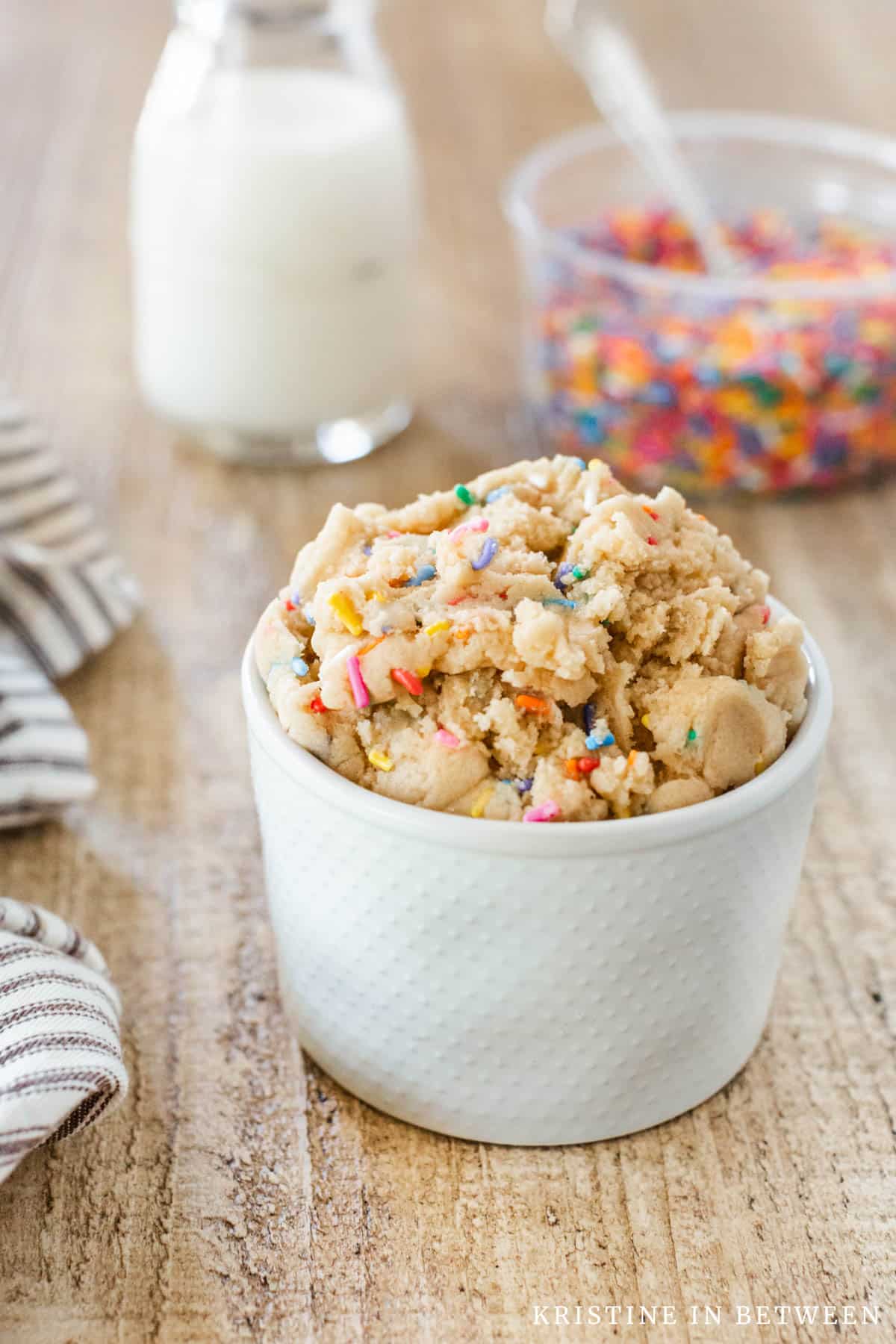 A small bowl of edible sugar cookie dough with sprinkles and a jar of milk and more sprinkles in the background.