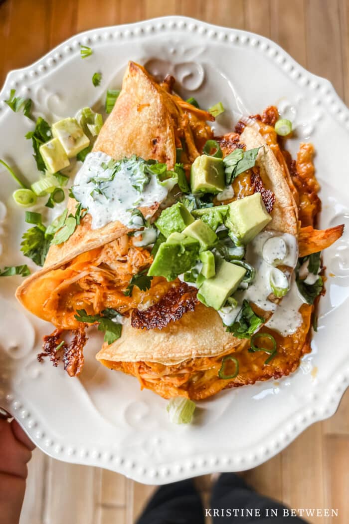 Two tacos sitting on a white plate with chopped avocado and cilantro on top.
