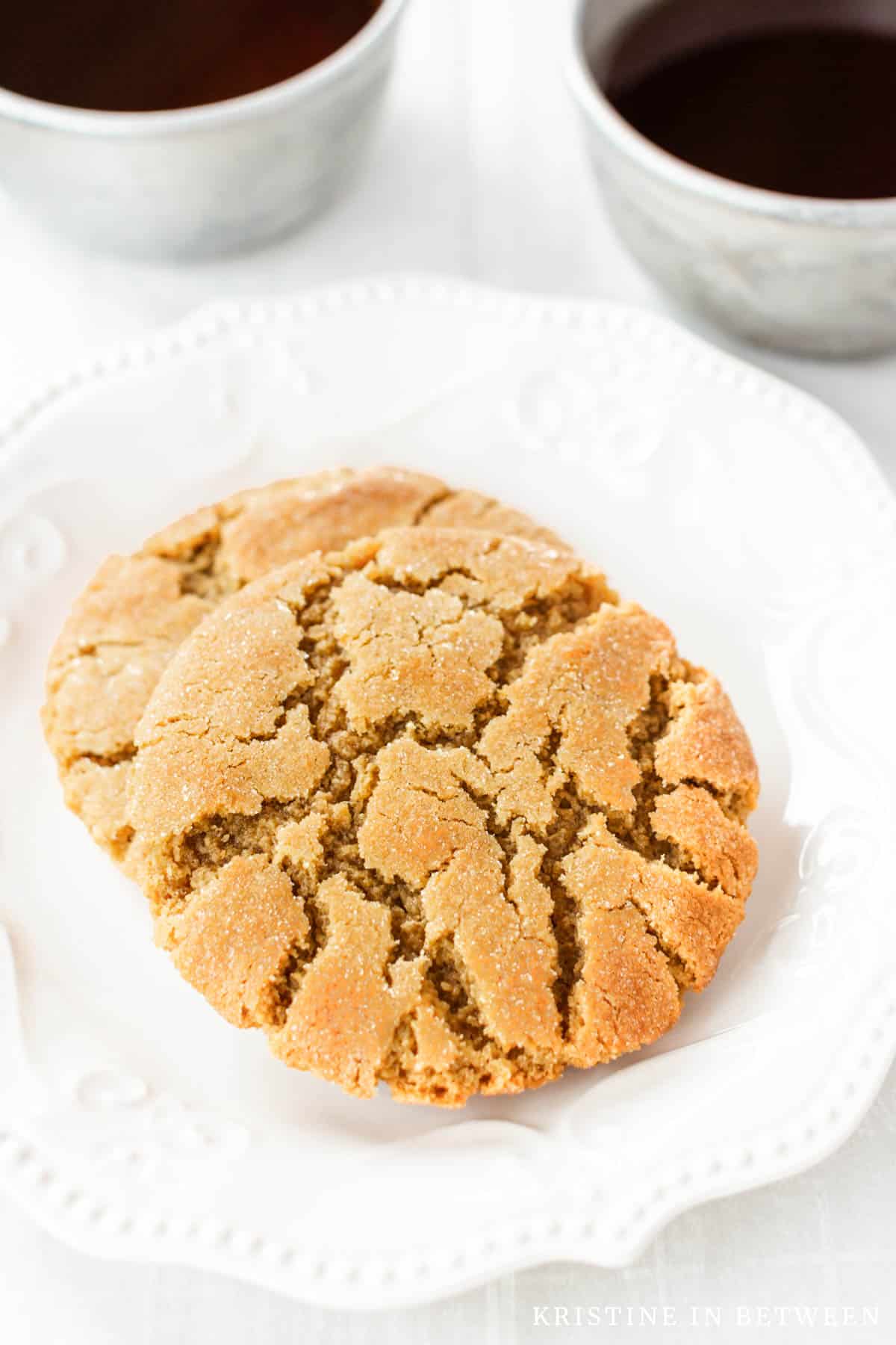 Two peanut butter cookies sitting on a white plate with coffee in the background.