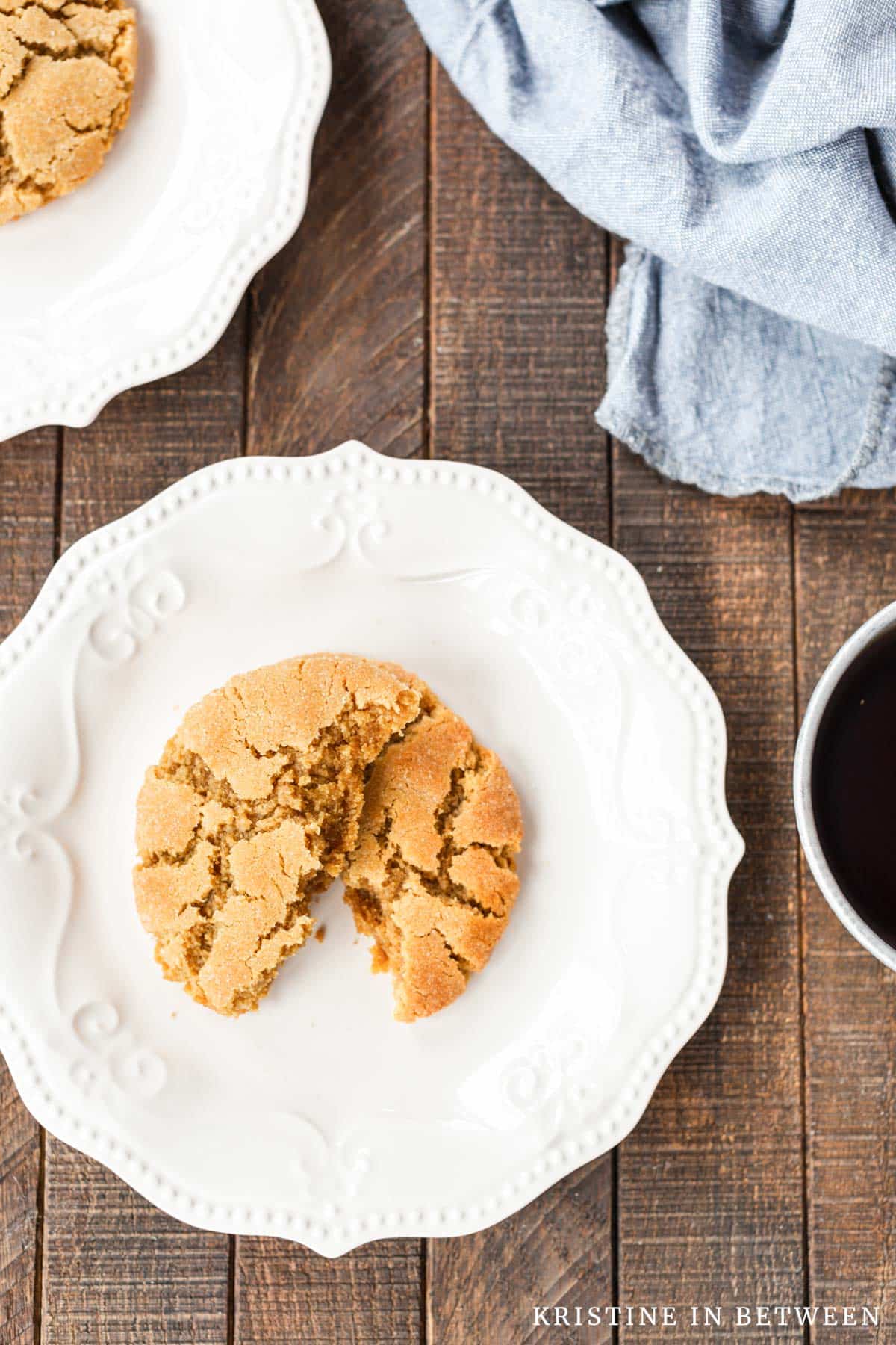 A peanut butter cookie that's broken in half on a white plate with a cup of coffee.