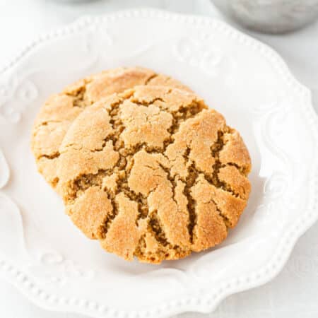 Two peanut butter cookies stacked up on a white plate with coffee in the background.
