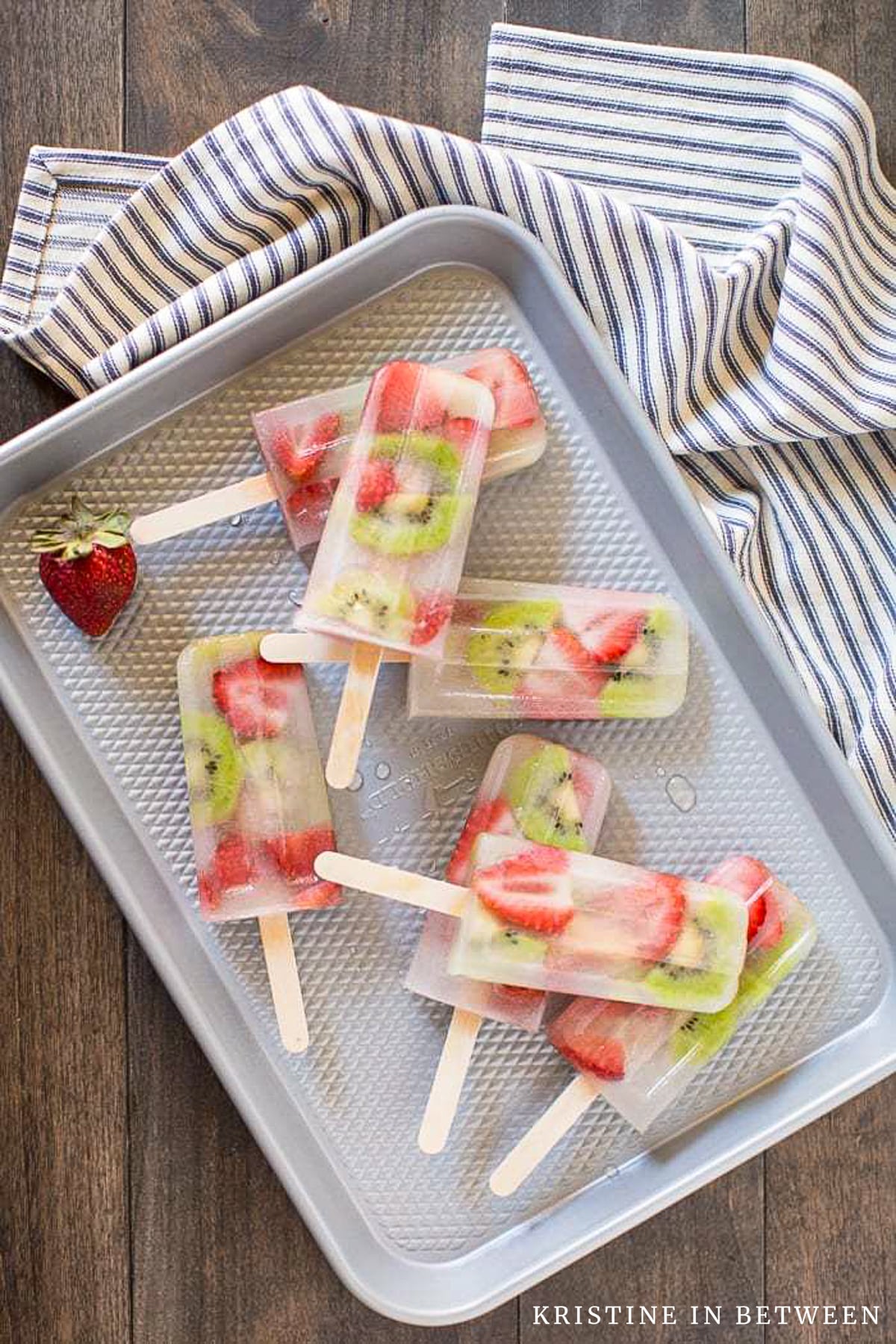Strawberry kiwi popsicles laying on a cookie sheet with a blue striped napkin next to them.