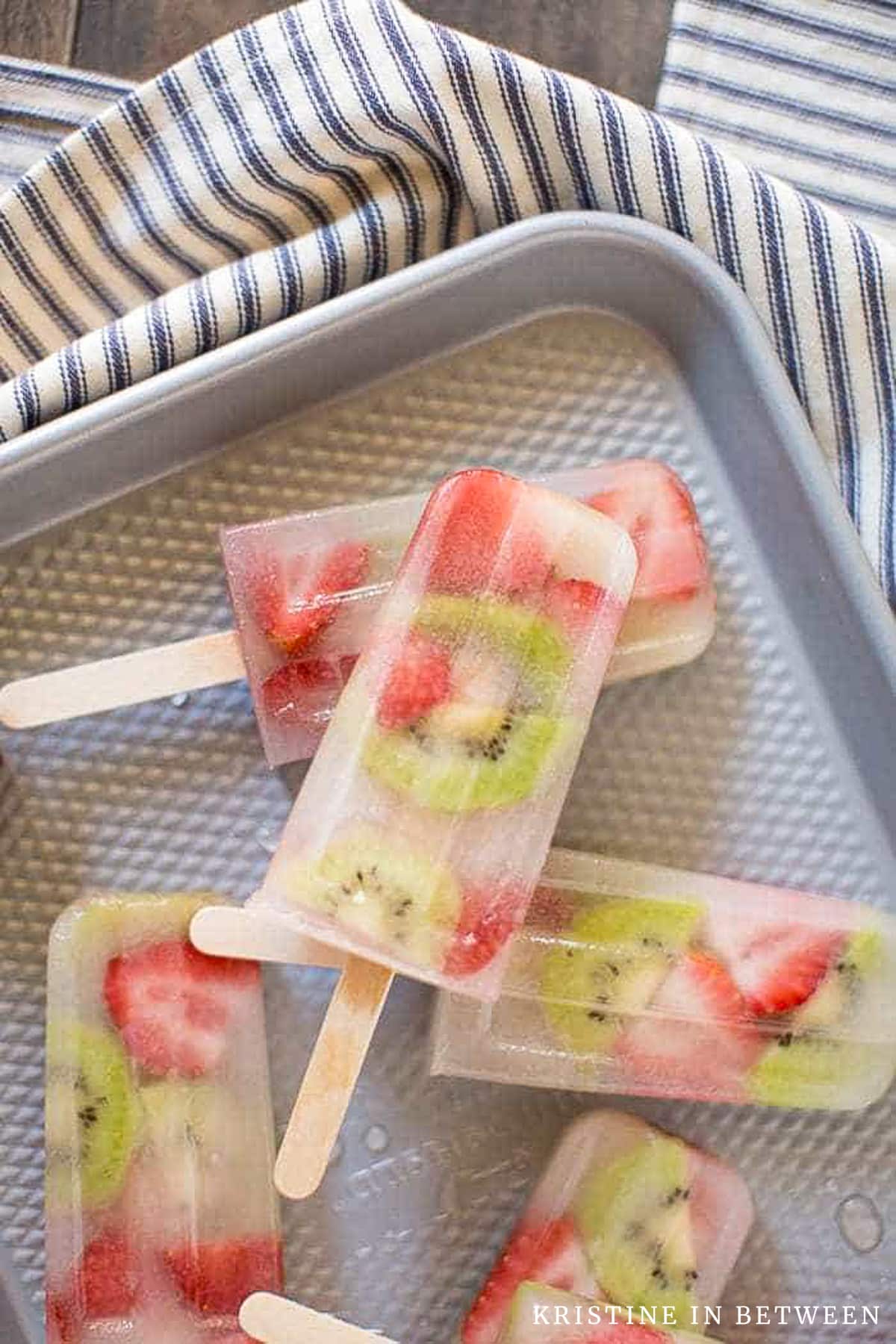 Strawberry kiwi popsicles laying on a cookie sheet with a blue striped napkin next to them.