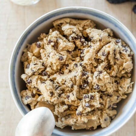 A bowl of edible chocolate chip cookie dough with a spoon it and a glass of milk in the background.