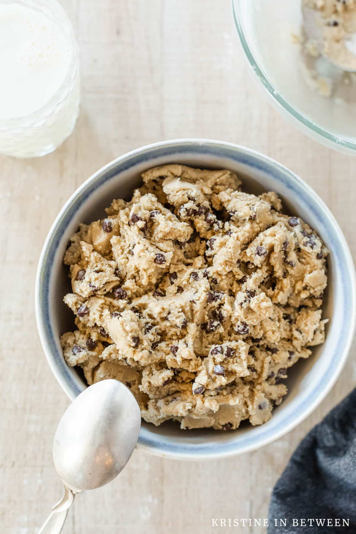 A big bowl of edible cookie dough with chocolate chips sitting with a spoon and a glass of milk.