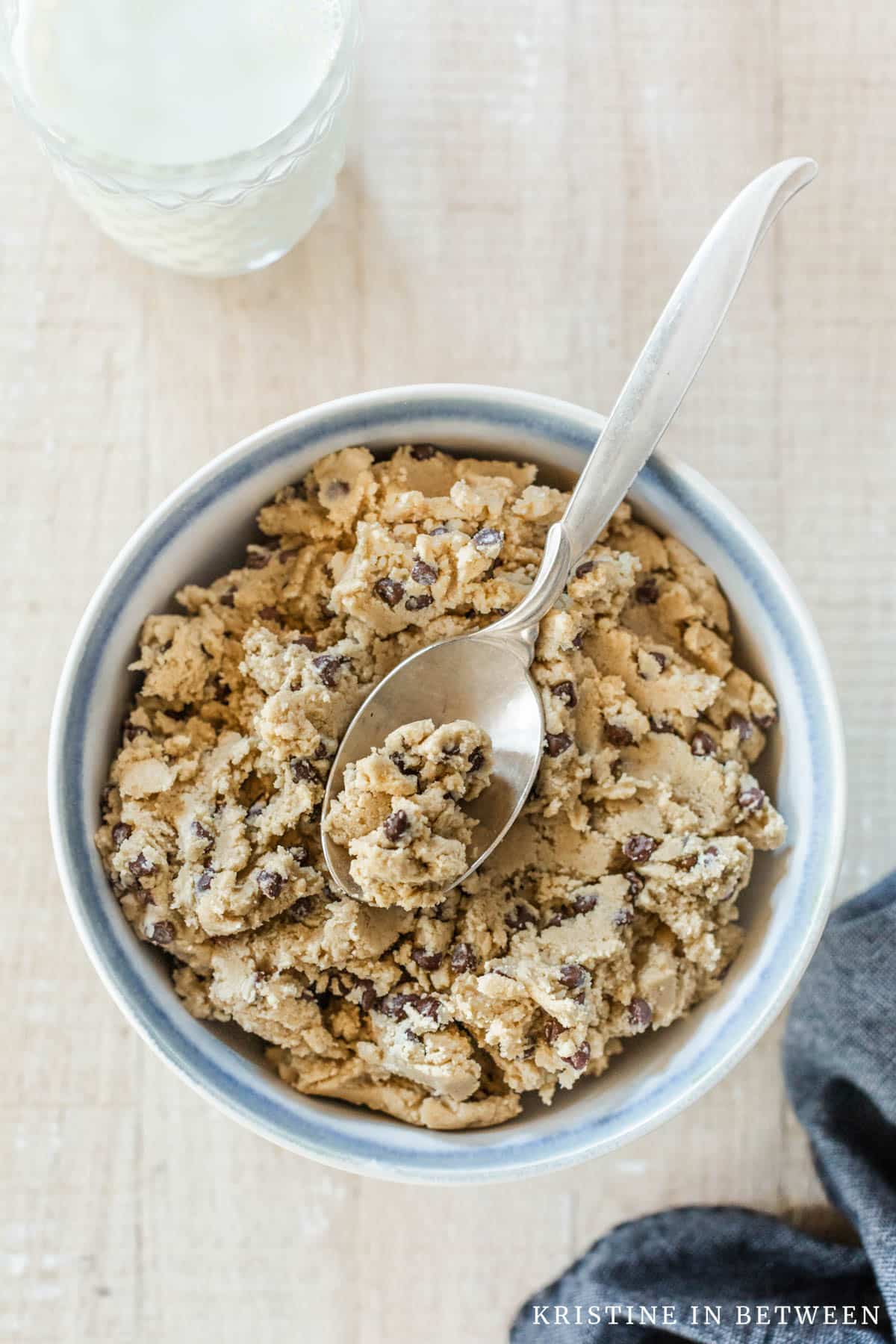 A bowl of edible chocolate chip cookie dough with a spoon it and a glass of milk in the background.