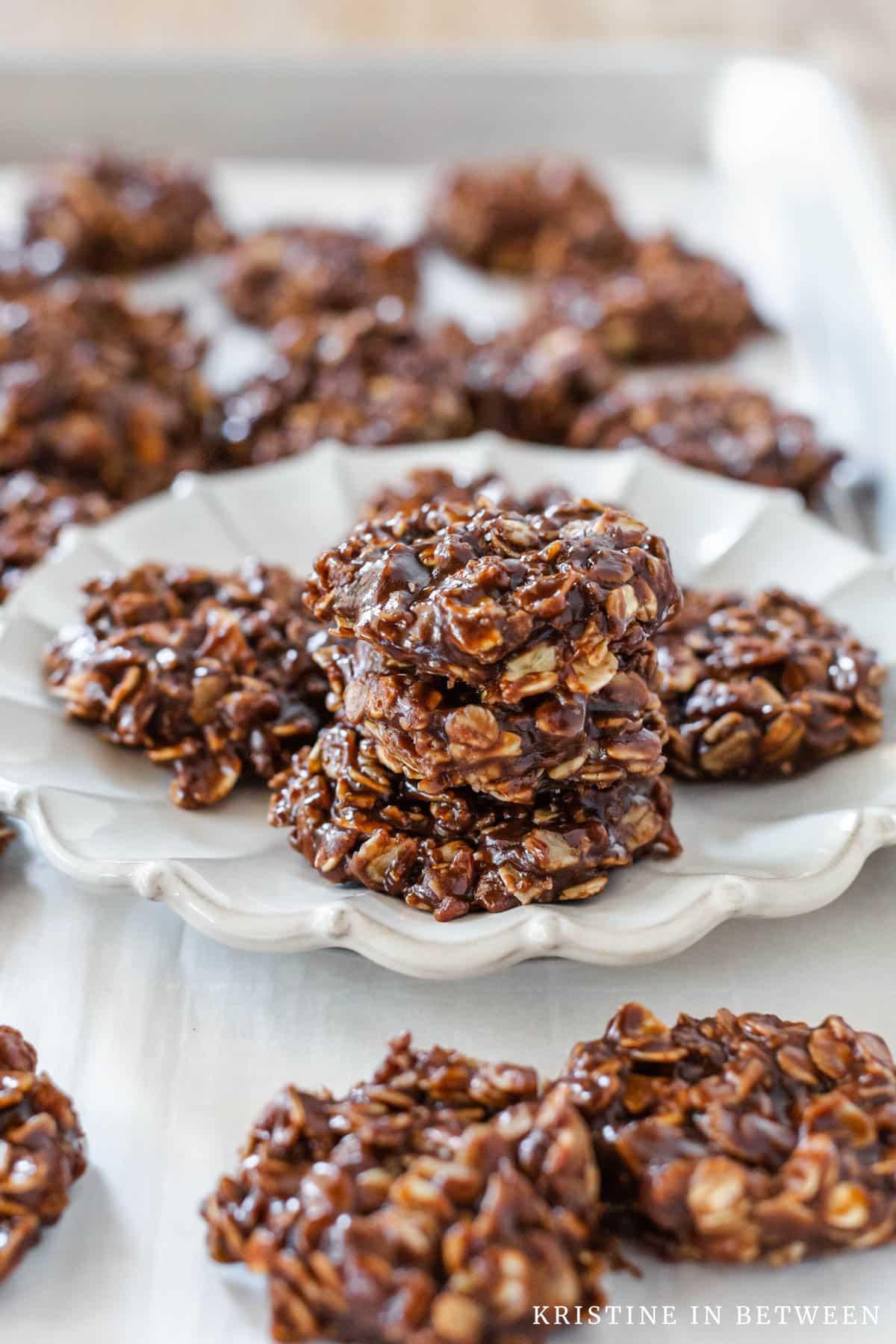 Several no-bake cookies stacked upon a white plate with more cookies in the background.