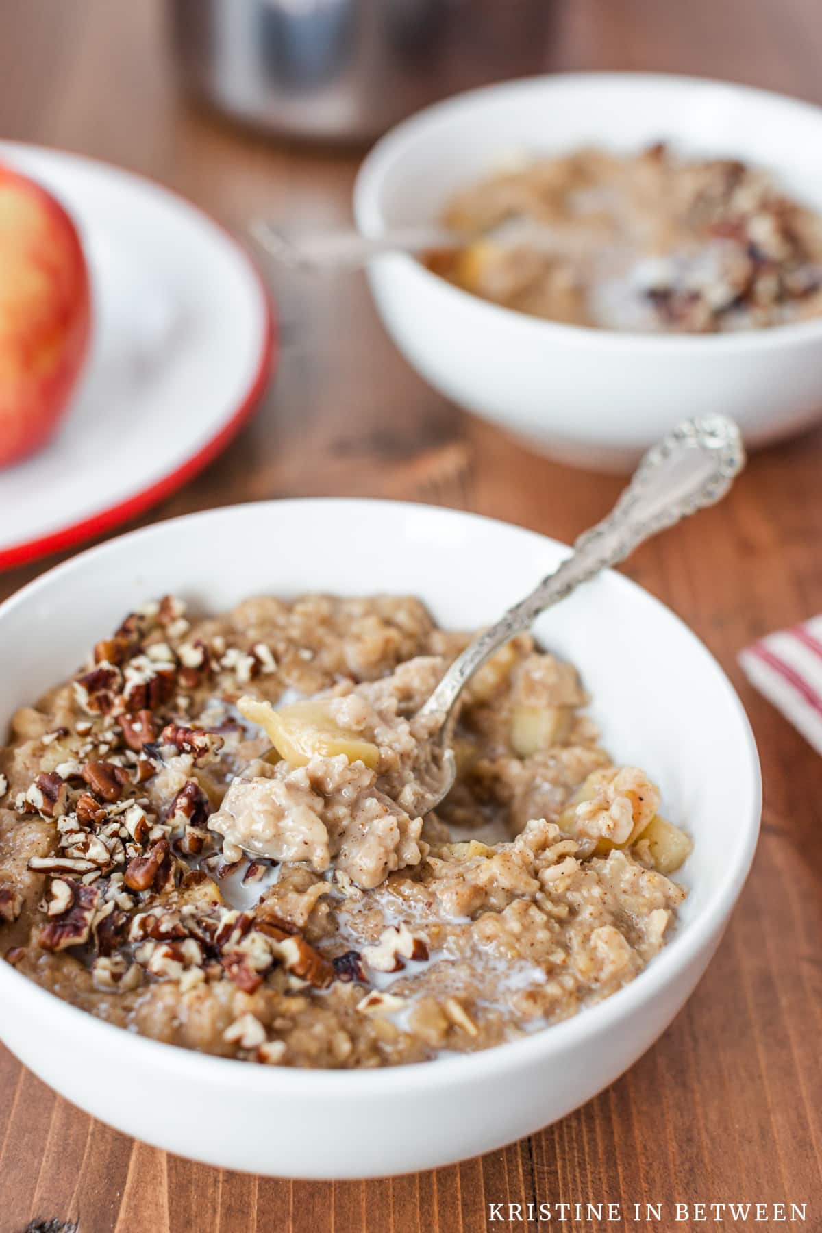 A bite of apple pie oatmeal on a spoon with another bowl and a pan in the background.