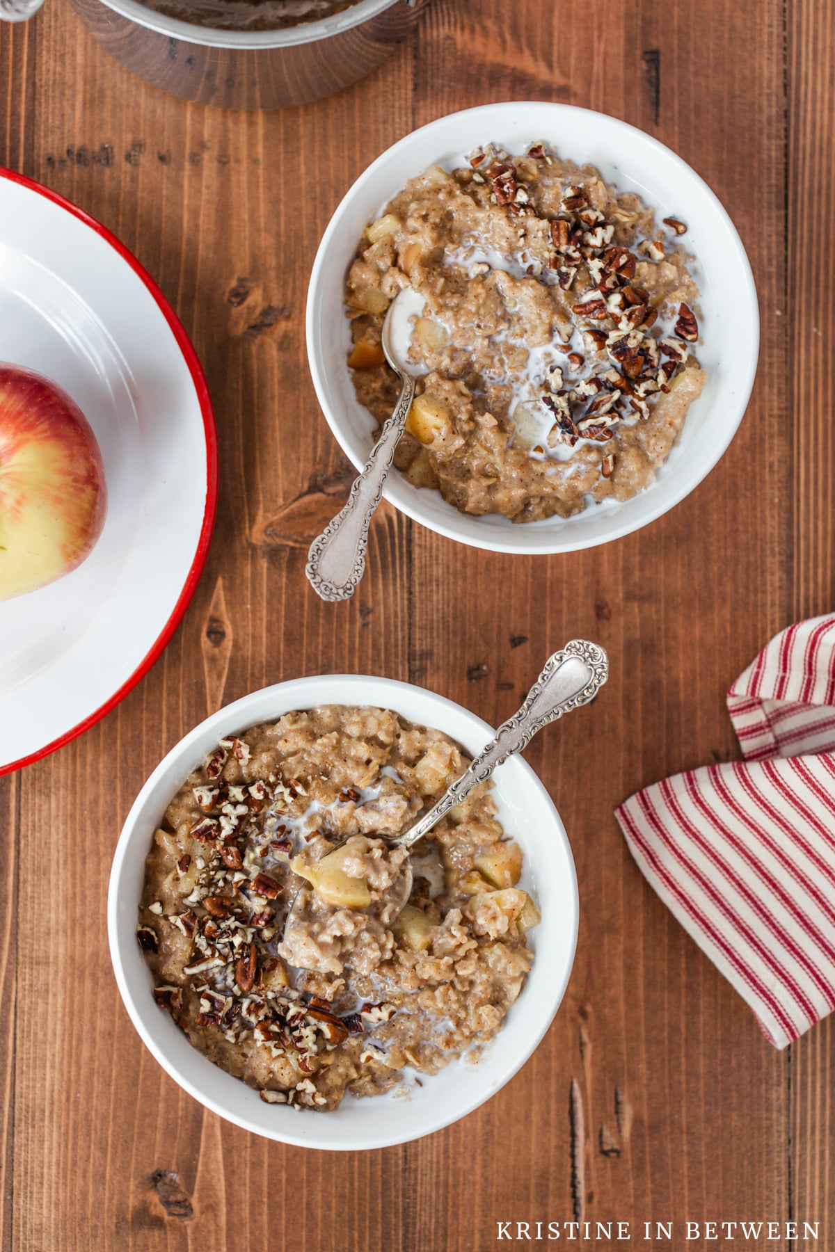 Two bowls of apple pie oatmeal with spoons in them and an apple sitting next to them.